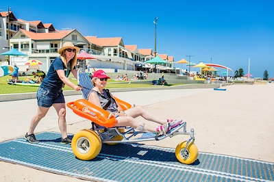 A woman is at the beach, seated in the Mobi-Chair Beach Wheelchair as its pushed by another woman along a stretch of blue beach access matting