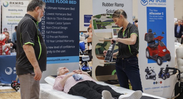 Stallholders demonstrate their products at the 2023 Seniors and Carers Expo. A hospital bed and mobility scooters are on show.