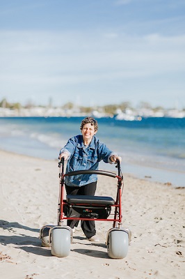 A smiling woman is in a blue shirt and black pants, walking along the beach using a walker with larger than usual wheels. This is a beach walker designed to help people move on the beach.