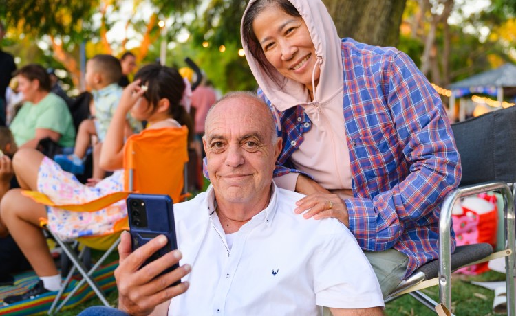 An older man and woman are smiling at the camera, the man is using a smart phone and is holding it in front of him
