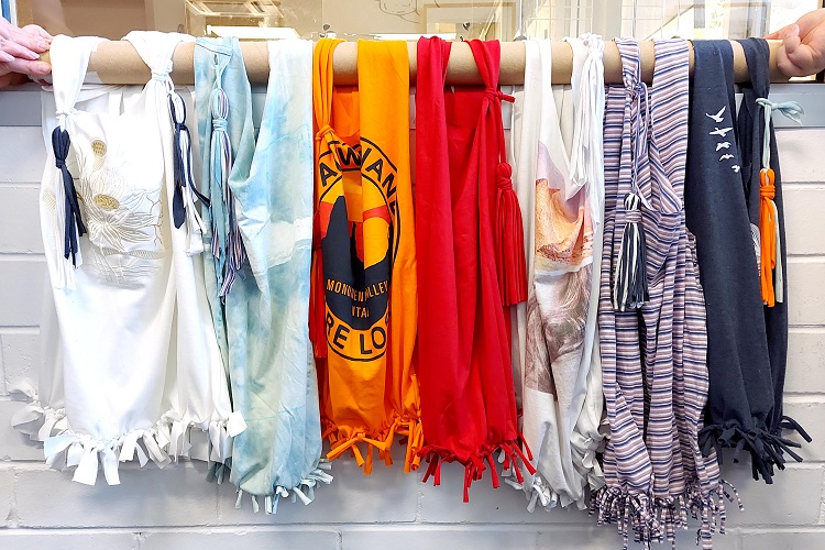 Seven tote bags made out of t-shirts hanging from a horizontal pole.
