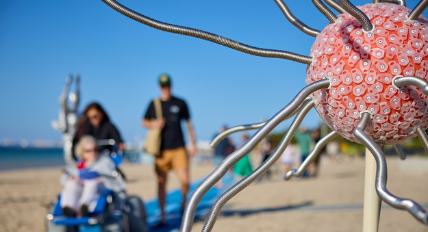 A spherical sculpture with metallic tendrils, made from a shark net float, contact lens containers and galvanized steel, displayed on the beach.