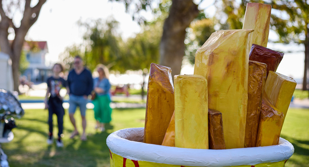 A sculpture of a giant bucket of chips, made from copper pipe, pine, cardboard, and canvas, displayed in a park with people in the background.