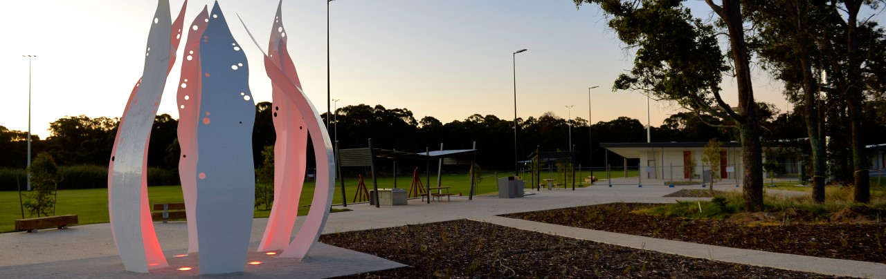 Outide at the Baldivis Indoor Sports Complex, the image shows the art sculpture, bench seats and tables under shades and lighting that surrounds one of the club house buildings infront of a playing field.