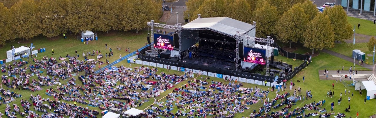 Ariel shot of a crowd on a large green space, seated on the ground in front of a stage. Screens on the stage are displaying the words 'Symphony on the Green'