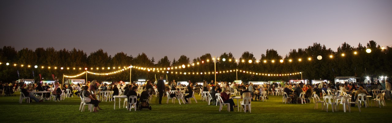 A crowd of people on a community oval at dusk. They are all seated around round tables and underneath string lights. In the background the light from food truck windows is also glowing