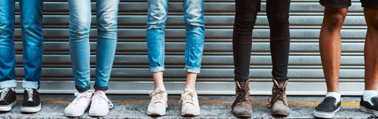 Image shows five pairs of legs standing infront of a corrugated iron door. All in jeans and casual shoes