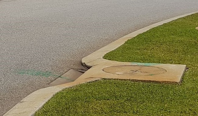 Concrete soakwell cover integrated into a grass verge next to a kerb inlet on a sealed road, with green survey markings on the kerb.