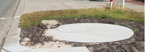 Circular concrete soakwell cover installed in a verge area with exposed soil and adjacent kerb, near a paved footpath and road.