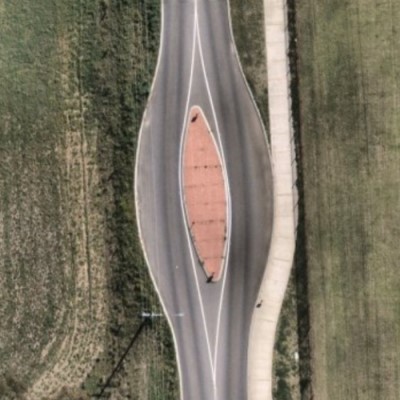 Aerial view of a road featuring a blister island—a raised, elongated, leaf-shaped median with reddish-brown brick or tile infill. The road curves around the island, with green fields and a narrow concrete strip visible alongside.
