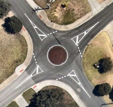 Aerial view of a mini roundabout with a central red‑coloured circular island, surrounding road markings, pedestrian kerb ramps, and adjacent landscaped areas.