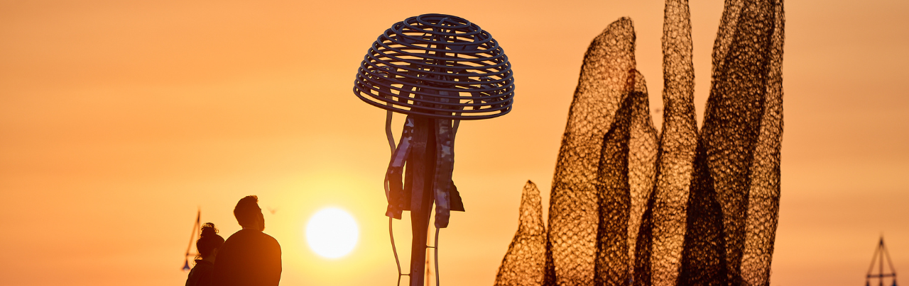 Two people look at an aluminium jelly fish sculpture and an abstract wire sculpture on the beach at sunset