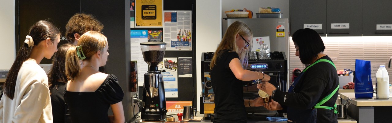 Standing infornt of a coffee machine, a young girl is pouring a cofee into a mug with the supervision and help from a older woman in a green apron. Gour oter studet aged people are standing and watching.