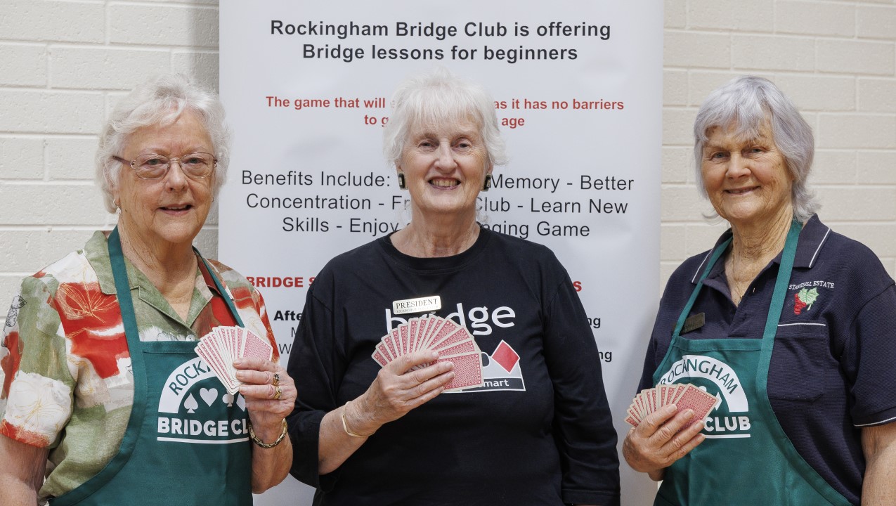 Three women are smiling at the camera all holding a hand of cards and standing infront of a sign that reads "Rockingham Bridge Club is offering Bridge lessons for beginners. The woman in the centre has a black Bridge Club shirt and the two other women are wearing green Bridge Club aprons
