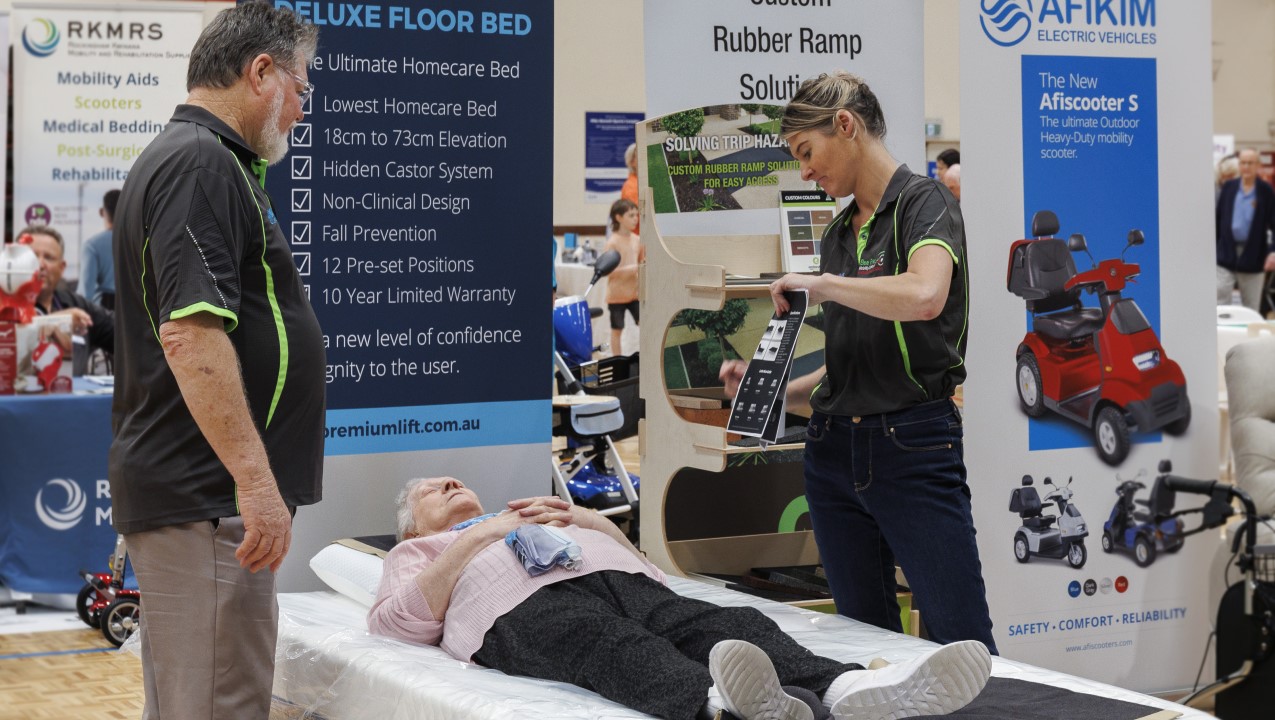 One person is lying on a bed for a product demonstration from the two people speaking to her. There are banners behind them advertising mobility products such as beds and scooters.