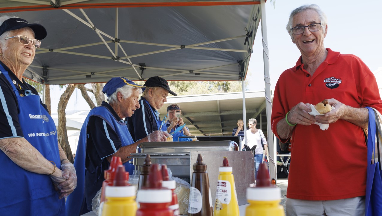 A man in a red shirt holds his sausage sizzle and smiles to the camera. Four people in Lions club aprons are working to cook more sausages as two more people are approaching in the background