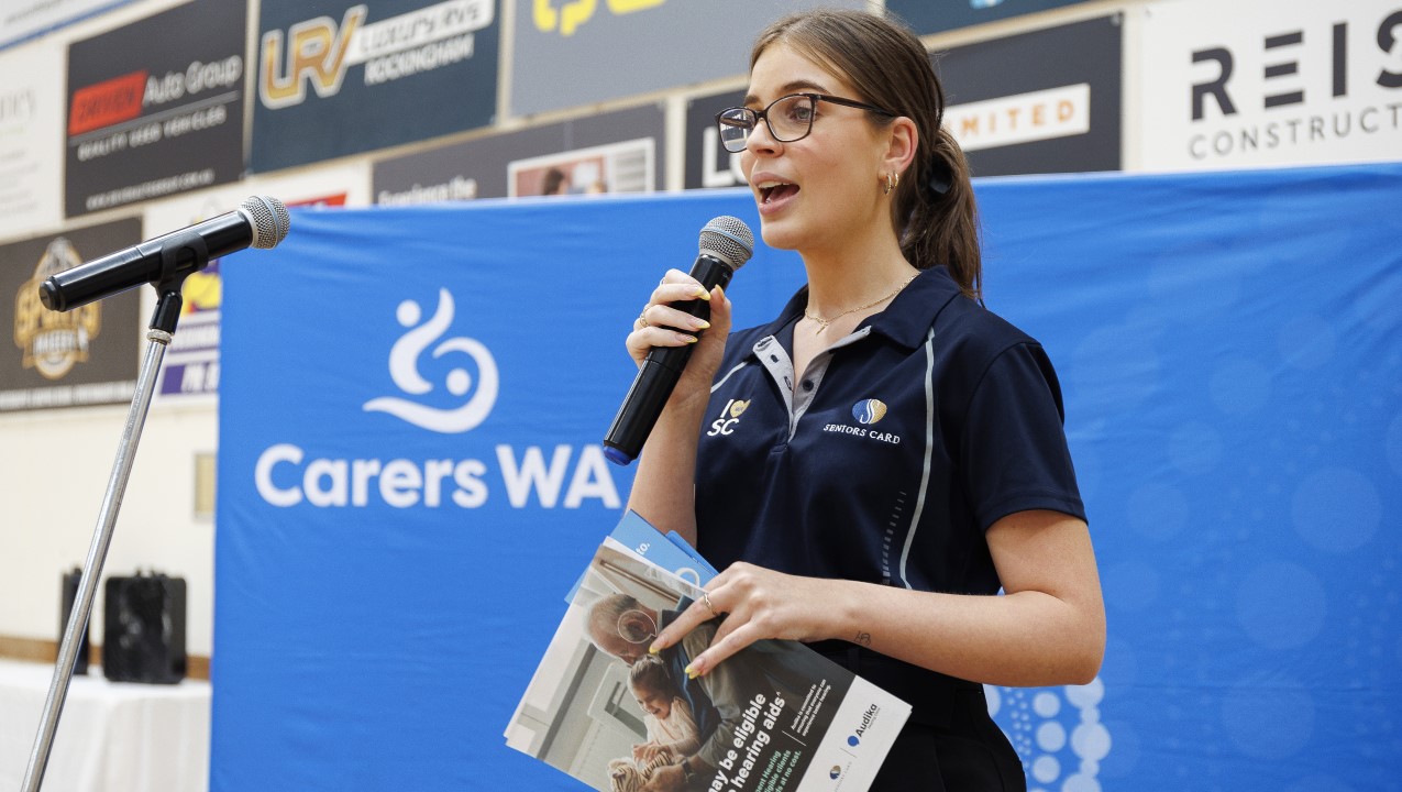 Woman wearing glasses, holding a brochure and microphone is standing in front of a blue Carers WA sign to give a speech.