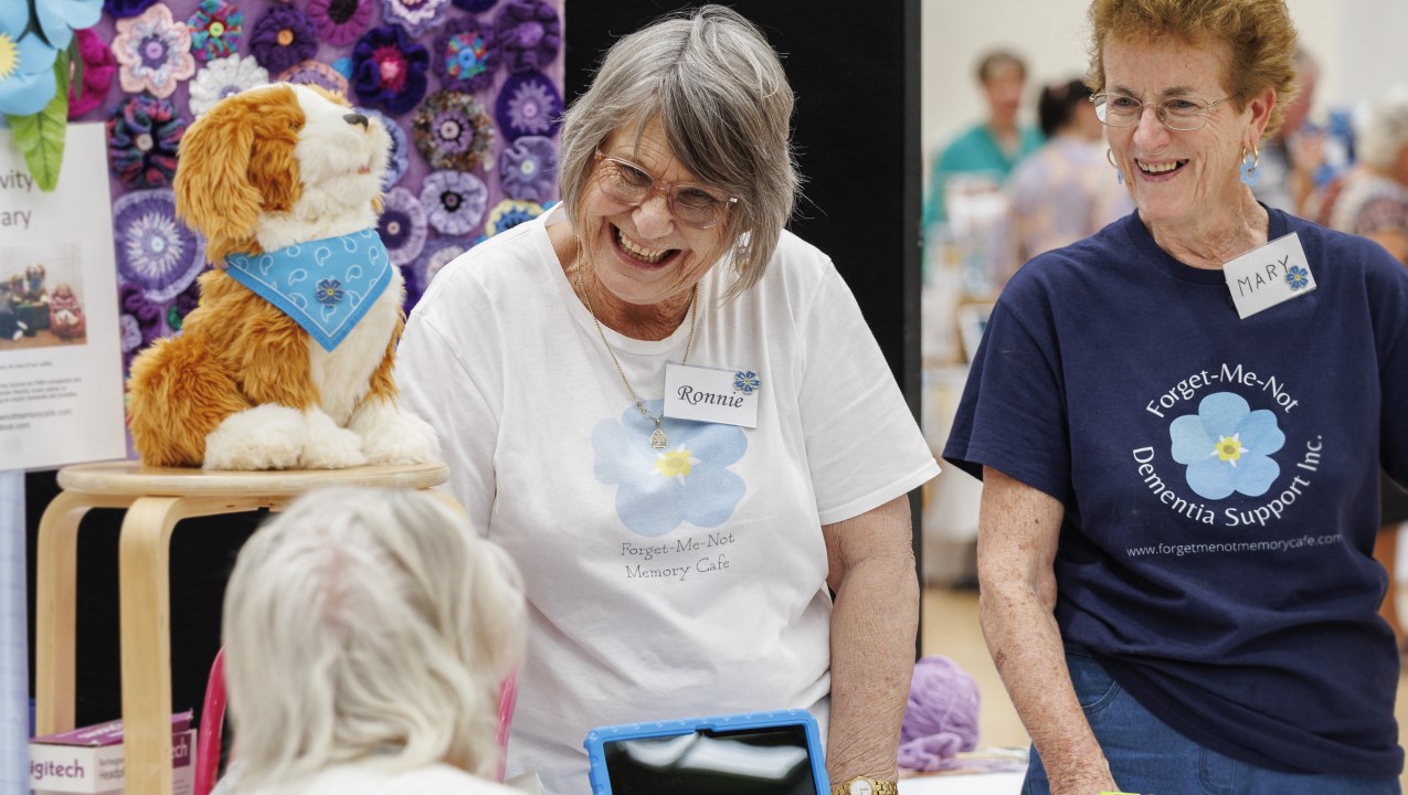 Two women are smiling and laughing as they talk to a third person not facing the camera. This happy chat is at the Forget Me Not stall with their toy dog and a purple crochet flower wall behind them.