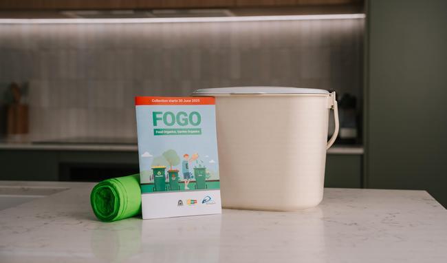 A FOGO guide is placed next to a kitchen caddy and compostable caddy liners on a kitchen bench.