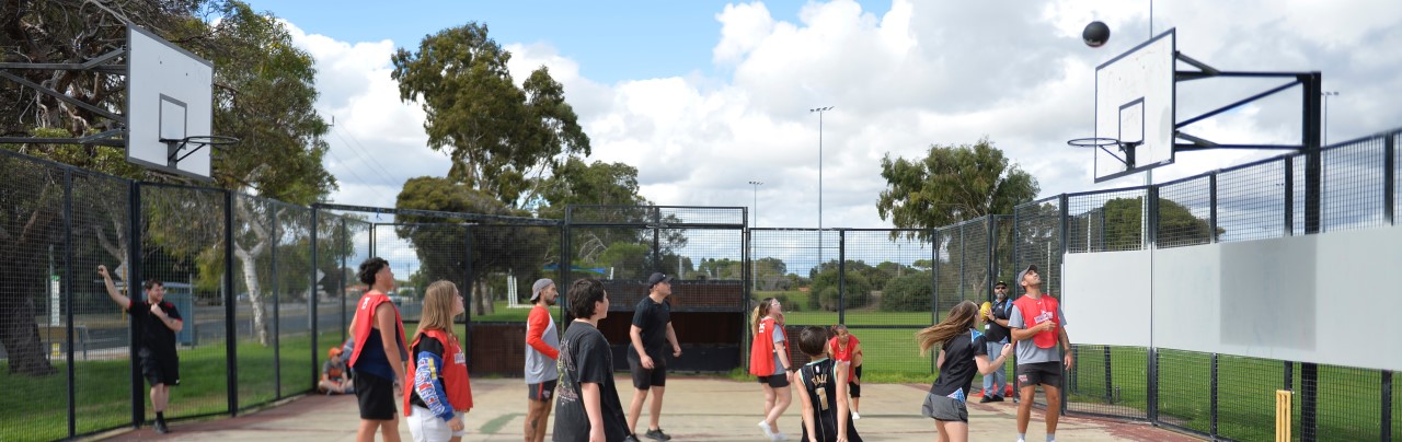 Ten people are arranged on an outdoor, enclosed by gates, basketball court. All ten players and the two people off court are watching a basketball approach the hoop after it was thrown by one of the players, with long hair and a black sirt