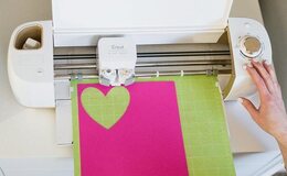 A hand operates a cricut machine while it cuts the outline of a heart on hot pink paper