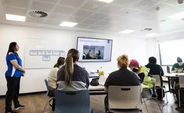 young people sat at tables looking towards a white screen with a presentation
