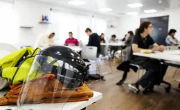 young people working at tables, a close up table with a high vis vest and protective face shield