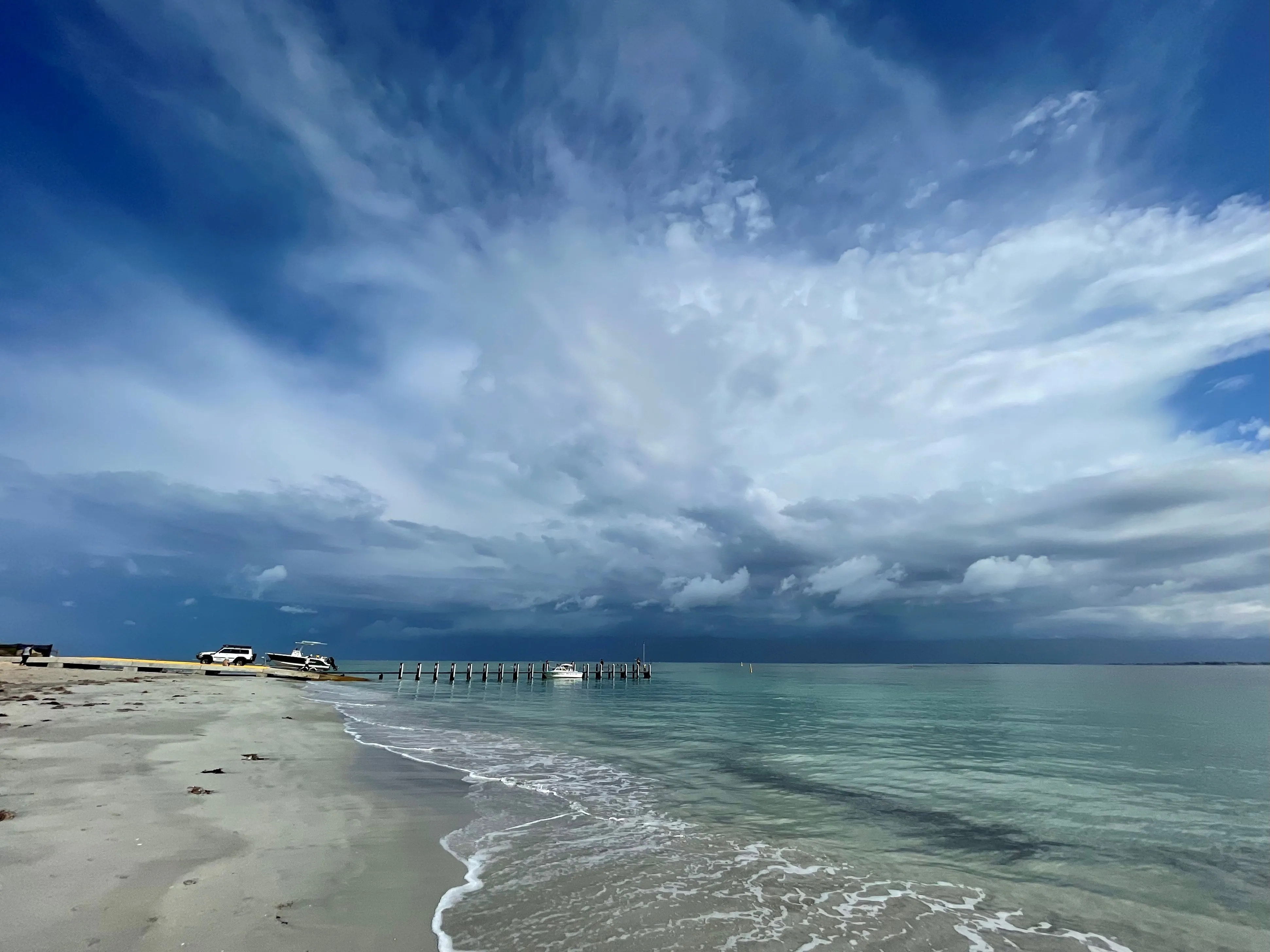 Port Kennedy Beach with pale sand, gentle turquoise waves, boats by a wooden pier, and dramatic layered clouds filling the sky.