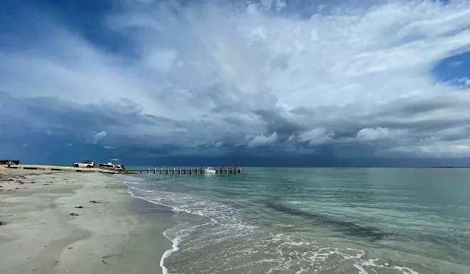 Port Kennedy Beach with pale sand, gentle turquoise waves, boats by a wooden pier, and dramatic layered clouds filling the sky.
