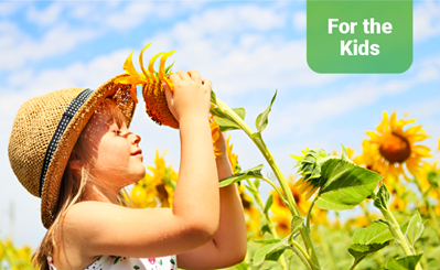 Child with their eyes closed, wearing a sun hat, face tilted towards the sun and holding a sunflower to their face.