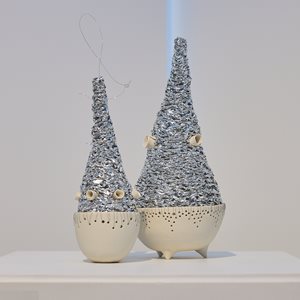 A sculpture of two abstract figures made from recycled high-fired porcelain and crinkled chip packaging. Each figure has a rounded ceramic base with delicate perforations and a textured metallic cone-shaped top, accented with small protruding elements.