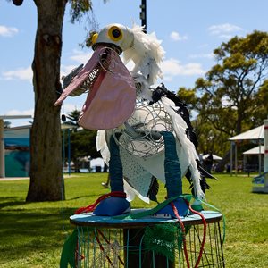 A sculpture of a pelican made from recycled materials, including a lampshade, wire fruit baskets, vacuum parts, milk bottles, tyre tubes, and a gym ball. The pelican appears to be eating discarded fishing lines and rubbish.